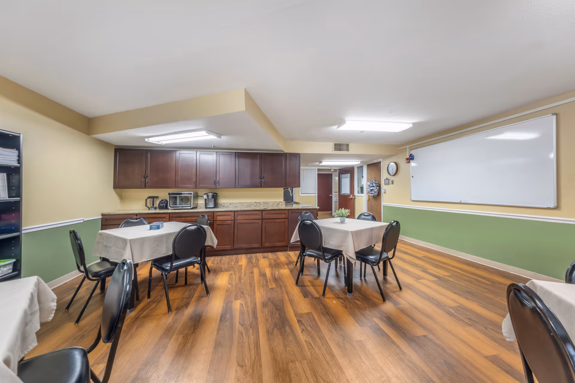 A communal room with several tables covered with beige tablecloths and black chairs around them. The room has wooden flooring, a green and beige painted wall, and a whiteboard mounted on the wall. There are dark wooden cabinets along one wall with a coffee maker, toaster oven, and other small appliances on the countertop. A clock and a wreath are visible on the far wall near a door.