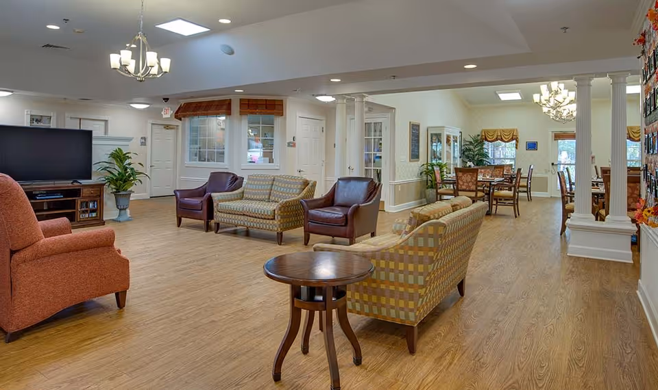 A spacious senior living common area with wooden flooring, multiple seating arrangements including armchairs and sofas, a large flat-screen TV on a wooden stand, and a dining area with tables and chairs in the background. The room is well-lit with ceiling lights and natural light from windows, decorated with plants and framed pictures.