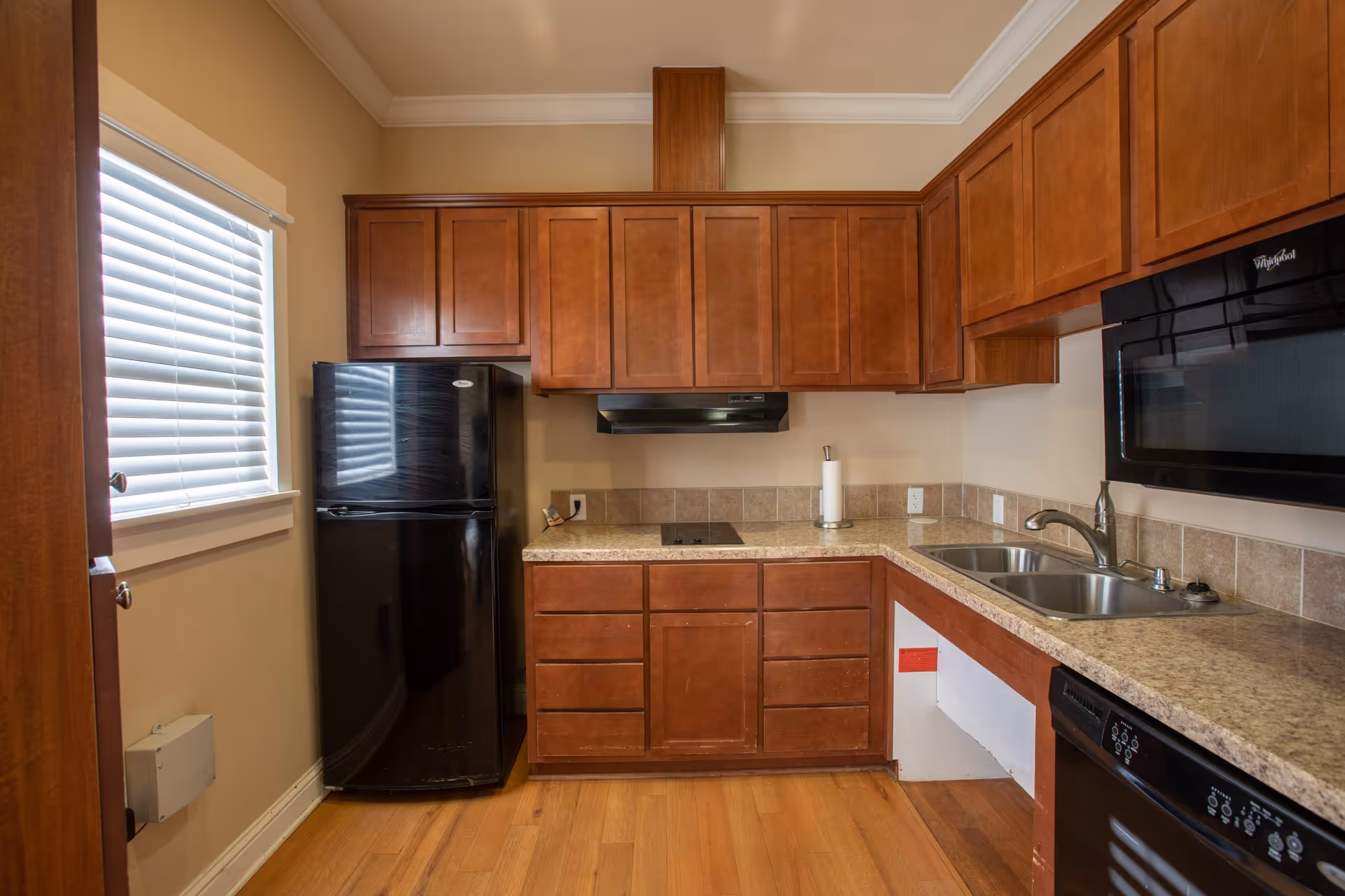 A kitchen with wooden cabinets, a black refrigerator, a black microwave, a black dishwasher, a double sink, and a countertop with a paper towel holder. There is a window with white blinds on the left side and wooden flooring.