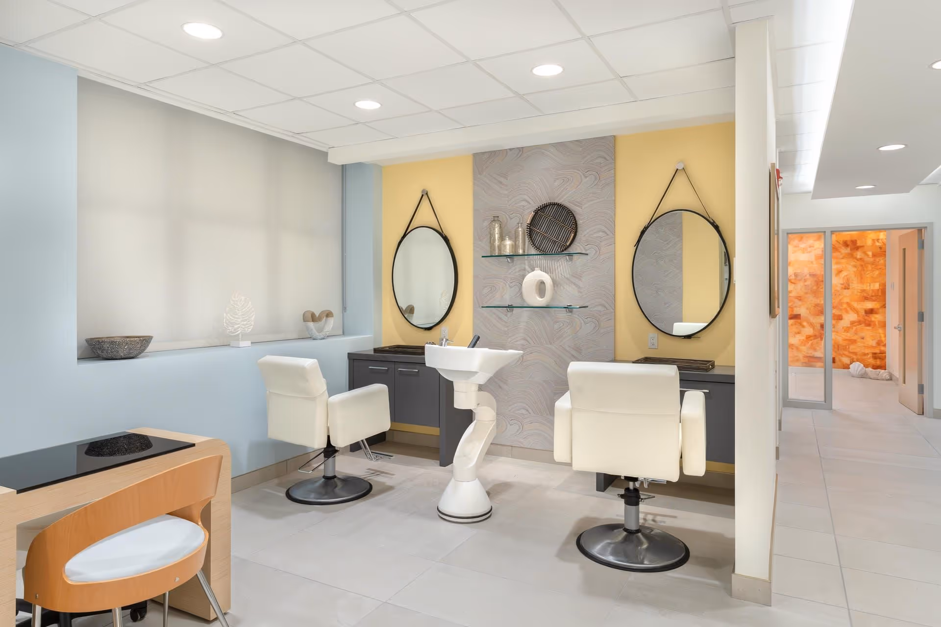 A bright salon-style interior with two styling chairs, round mirrors, a shampoo basin, and a small manicure table.
