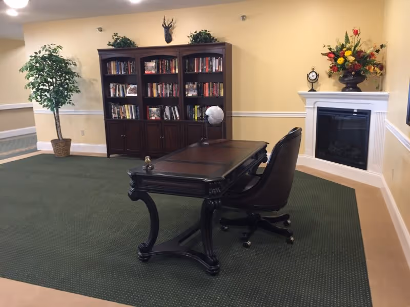 A cozy interior room with a dark wooden desk and black leather office chair on a green carpet. Behind the desk is a large wooden bookshelf filled with books and decorative items. To the right, there is a white fireplace with a clock and a vase of colorful flowers on the mantel. A potted plant is placed near the left wall, and the walls are painted light yellow with white trim.