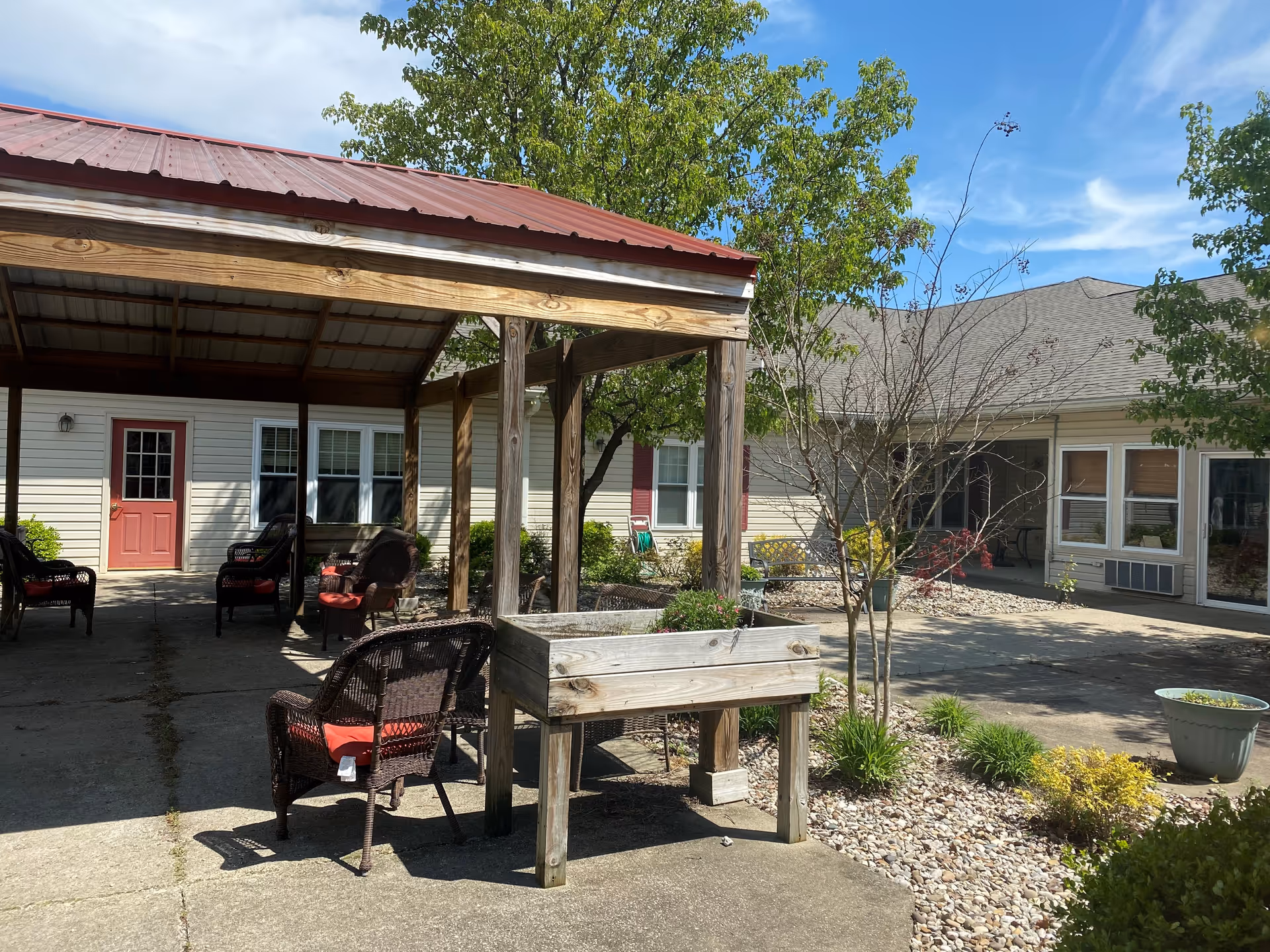 Outdoor courtyard area with a covered seating space featuring wicker chairs with red cushions. There is a wooden planter box with some plants, surrounded by small trees, shrubs, and a rock garden. The building exterior has beige siding with red doors and windows, under a clear blue sky.