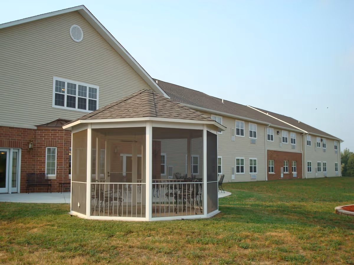 Outdoor view of a senior living facility named Heritage Woods of Mt. Vernon showing a large two-story building with beige siding and brick accents. In the foreground, there is a white screened gazebo with a shingled roof and outdoor seating inside. The area around the building is grassy with a clear sky above.