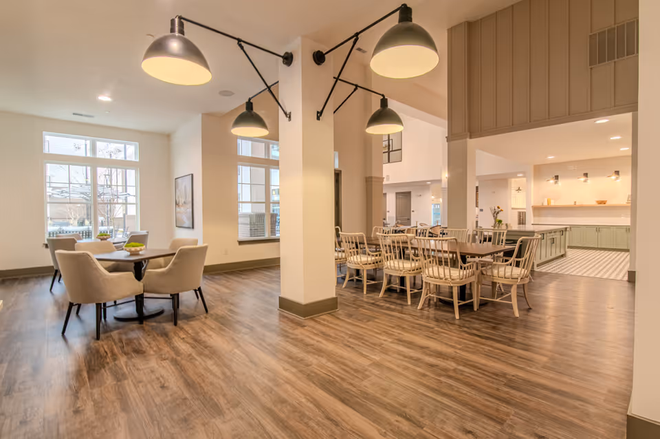 Bright open communal dining and seating area with wooden floors, multiple tables and hanging pendant lights near a kitchen counter.
