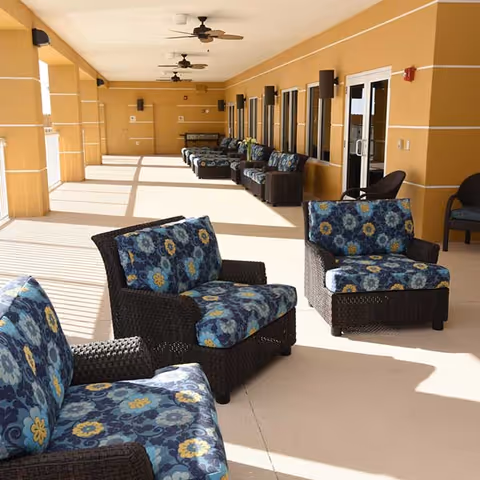 A spacious covered outdoor patio area with multiple cushioned wicker chairs featuring blue floral patterns. The patio has ceiling fans and is adjacent to a building with large windows and doors.