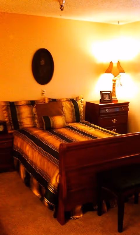 A warmly lit bedroom featuring a wooden sleigh bed with striped bedding, a nightstand with a lamp and framed photo, and a decorative wall plaque.