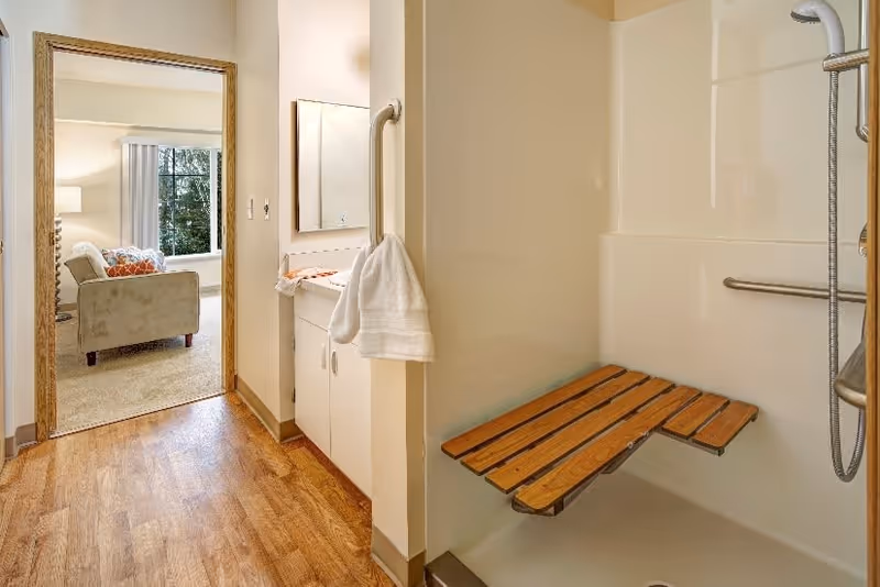 Interior view of a senior living facility bathroom with a walk-in shower featuring a wooden fold-down bench and grab bars. A towel hangs on a grab bar near a small sink and mirror. Through an open doorway, a living room with a beige armchair, a window with vertical blinds, and a lamp is visible.