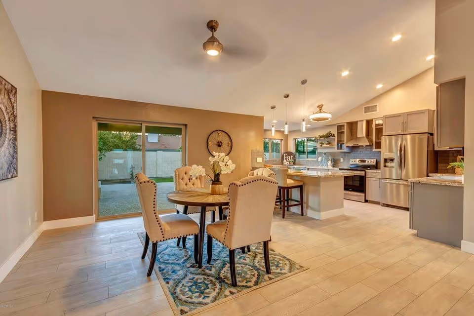Open-plan dining area with a round table and beige upholstered chairs adjacent to a modern kitchen and sliding glass door.