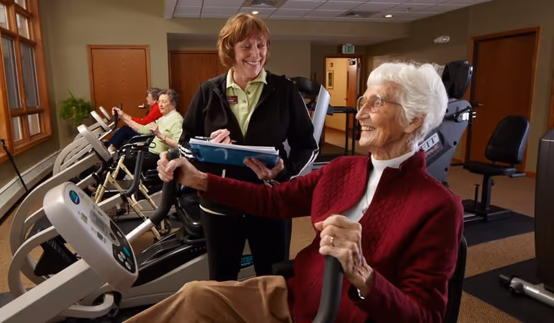 An elderly woman in a red sweater is using a stationary exercise bike in a fitness room at a retirement community. A staff member stands beside her, smiling and holding a clipboard. In the background, two other elderly women are also exercising on similar equipment. The room has beige walls, wooden doors, and exercise machines arranged neatly.