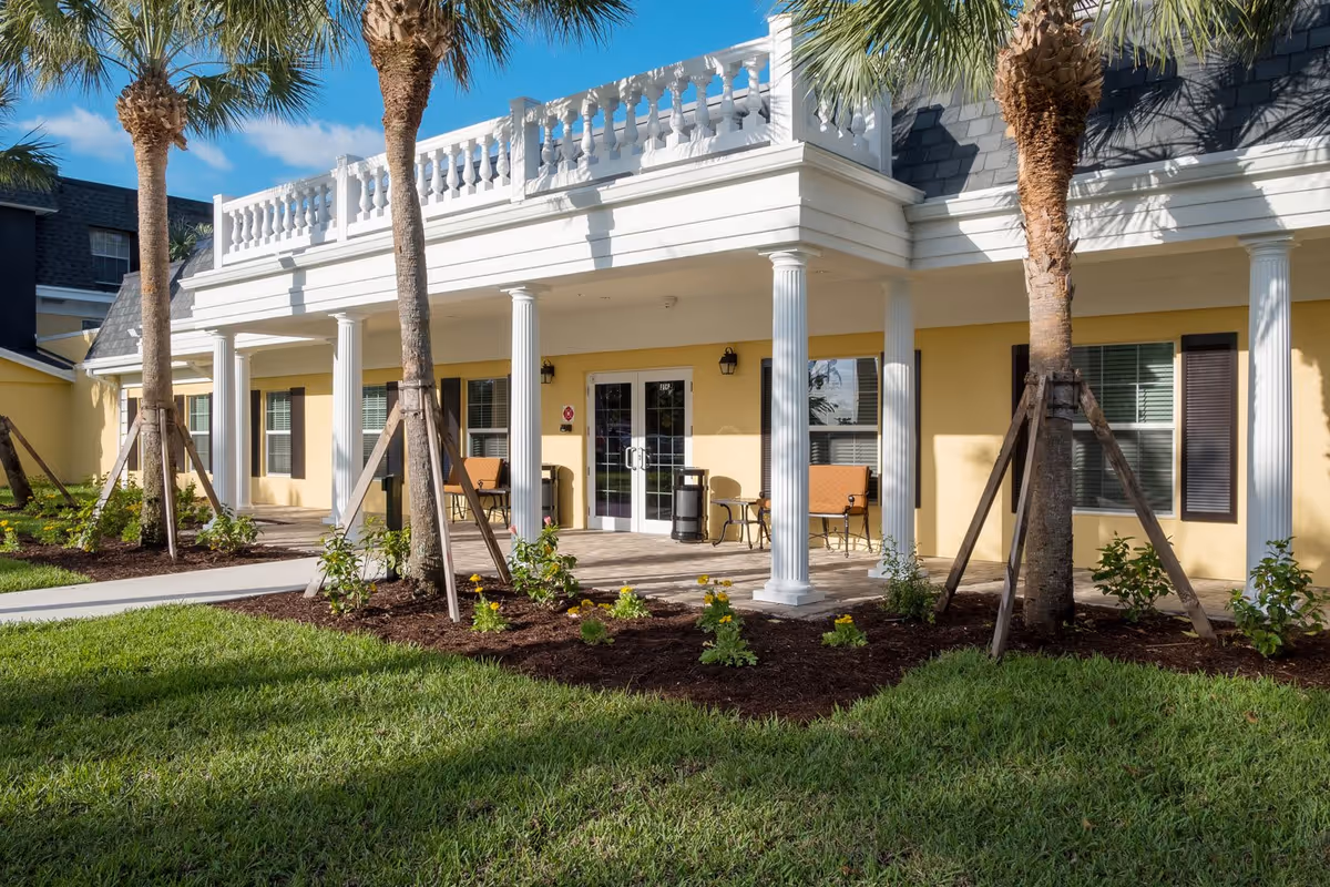 Exterior view of a senior living facility with yellow walls, white columns, and a balcony with white balustrades. There are palm trees and landscaped flower beds in front of the building, along with a paved walkway leading to double glass doors. Two benches and small tables are placed under the covered porch area.