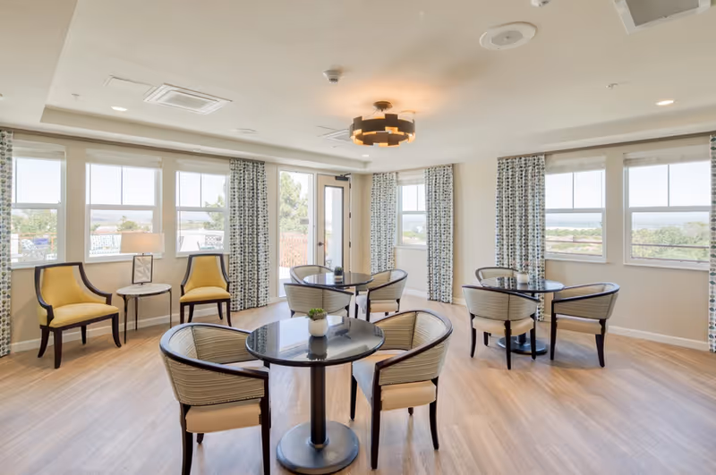 Bright communal dining/lounge area with round glass-top tables, upholstered chairs, and large windows with patterned curtains.