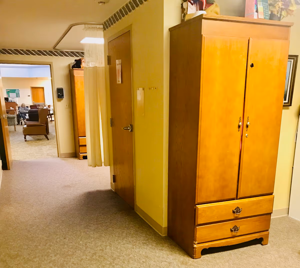 Interior hallway of a senior care facility with beige carpet and walls. A wooden wardrobe with two drawers is positioned on the right side. At the end of the hallway, there is a room with chairs and an elderly person sitting in a wheelchair. A curtain hangs from the ceiling near a door on the left side of the hallway.