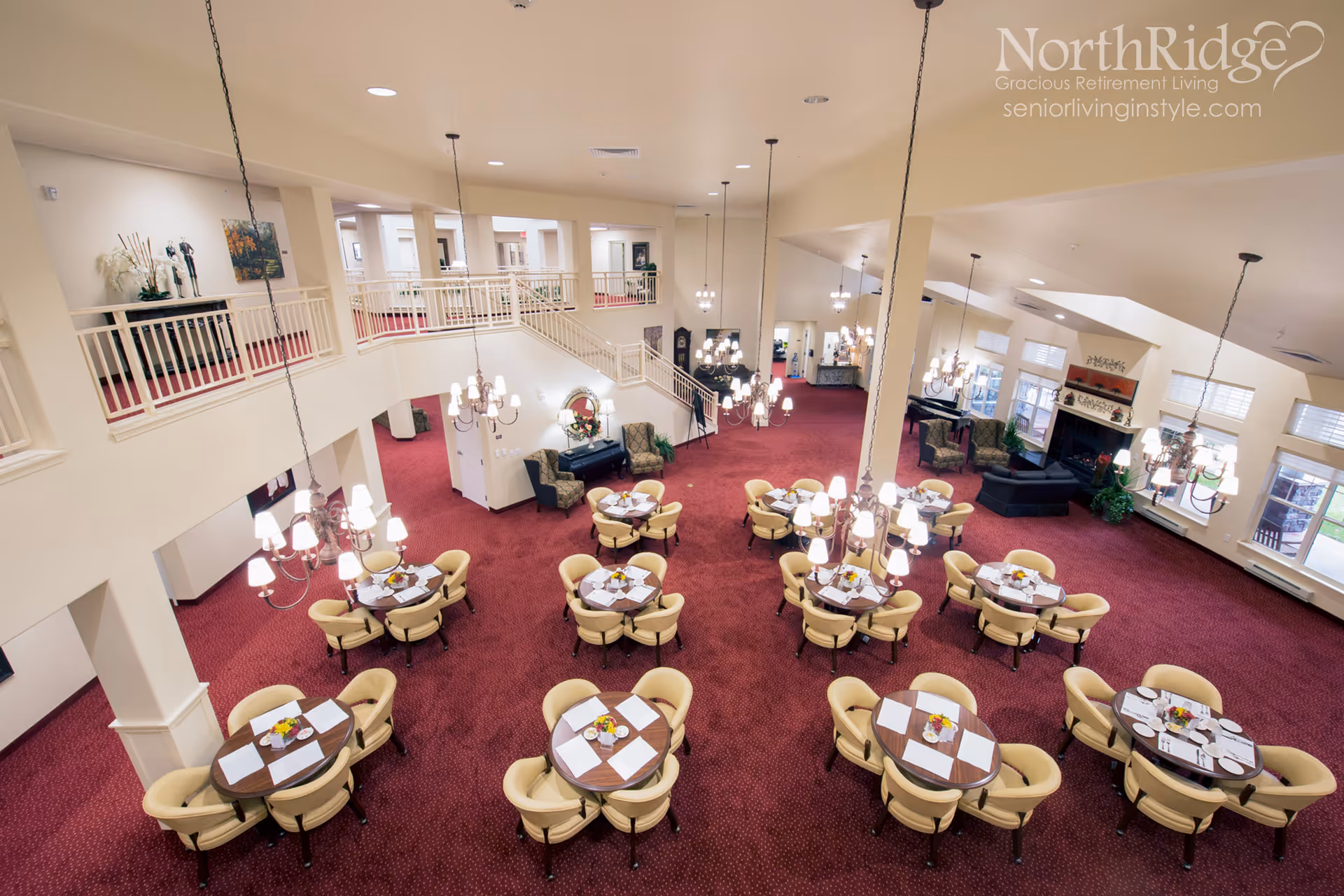 Large two-story retirement community dining and lounge area with round tables, beige chairs, chandeliers and red carpeted floor.