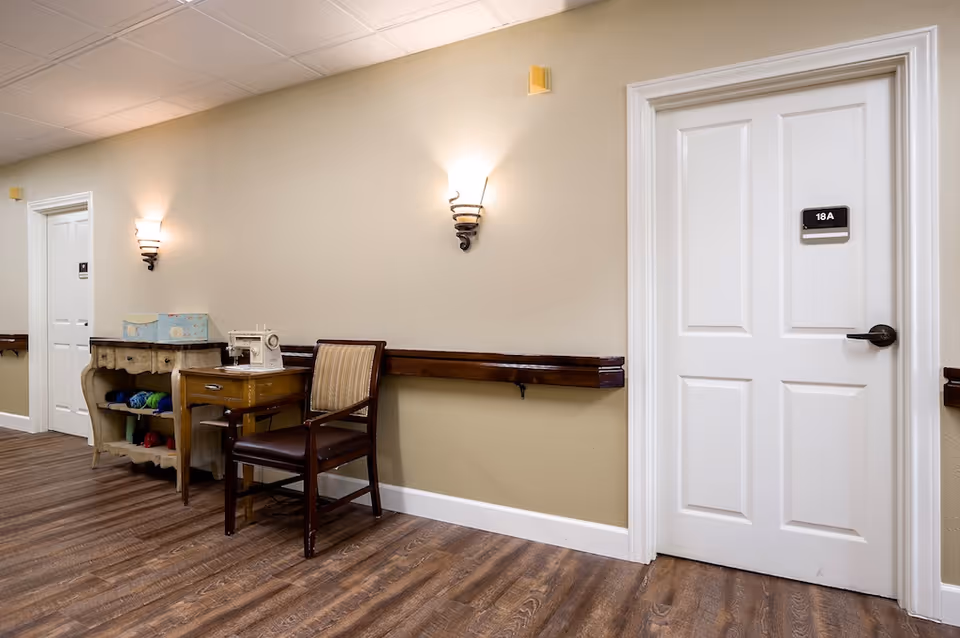 A hallway in a senior living facility with wooden flooring and beige walls. There are two white doors, one labeled 18A. The hallway features a wooden chair with striped upholstery and a small wooden table with a vintage sewing machine on top. Next to it is a small shelf holding colorful yarn. Wall-mounted light fixtures illuminate the space.