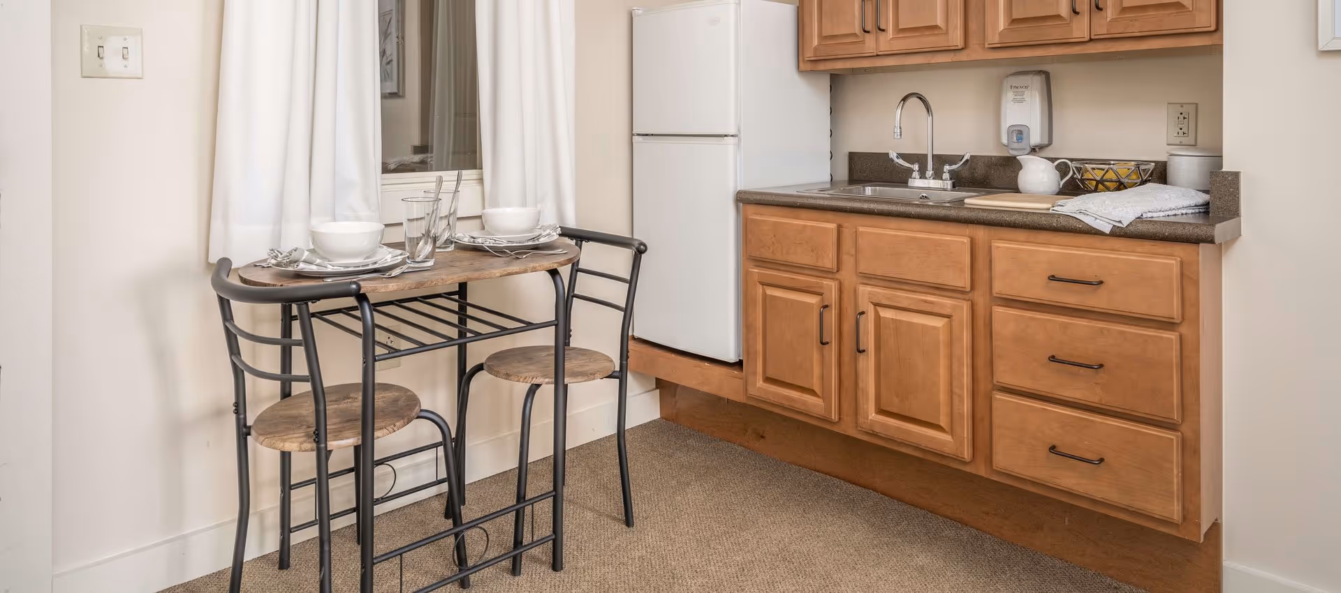 Small kitchen area with wooden cabinets, a white refrigerator, a sink, and a countertop with a pitcher, bowl of lemons, and a towel. Next to the kitchen is a small dining table set for two with plates, bowls, glasses, and utensils, positioned in front of a window with white curtains.