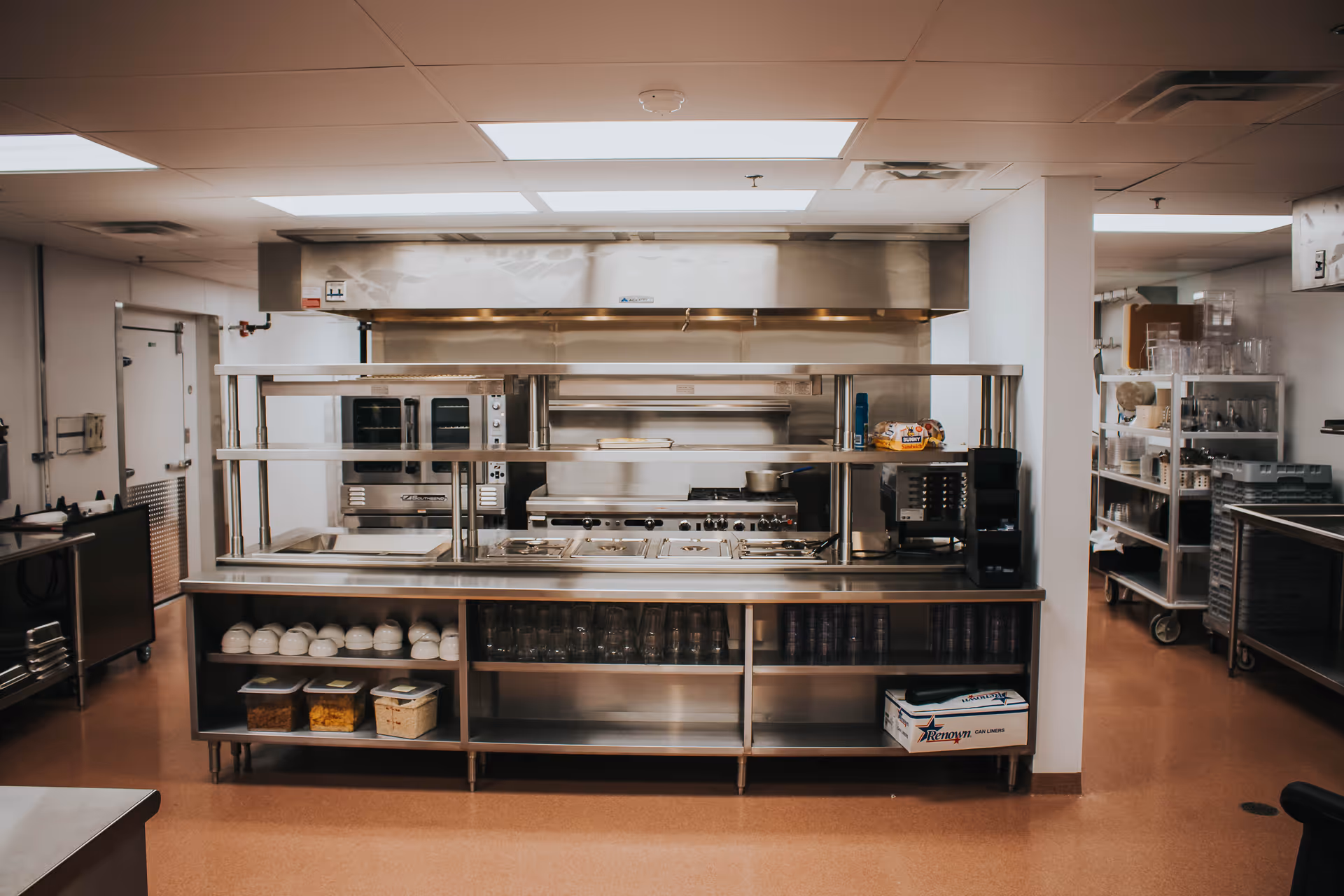 A clean and organized commercial kitchen with stainless steel counters and shelves. The kitchen features multiple ovens, a stove with pots, a coffee machine, and various kitchen supplies including bowls, glasses, and containers. The floor is a reddish-brown color and the ceiling has fluorescent lighting.