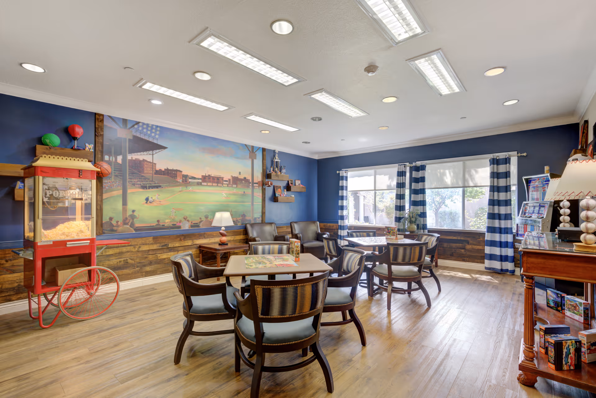 A bright and inviting common room with wooden floors, several tables and chairs arranged for group seating, a popcorn machine on the left, a mural of a baseball stadium on the blue wall, two armchairs near the window with striped curtains, and a wooden cabinet with games and a baseball-themed lamp.