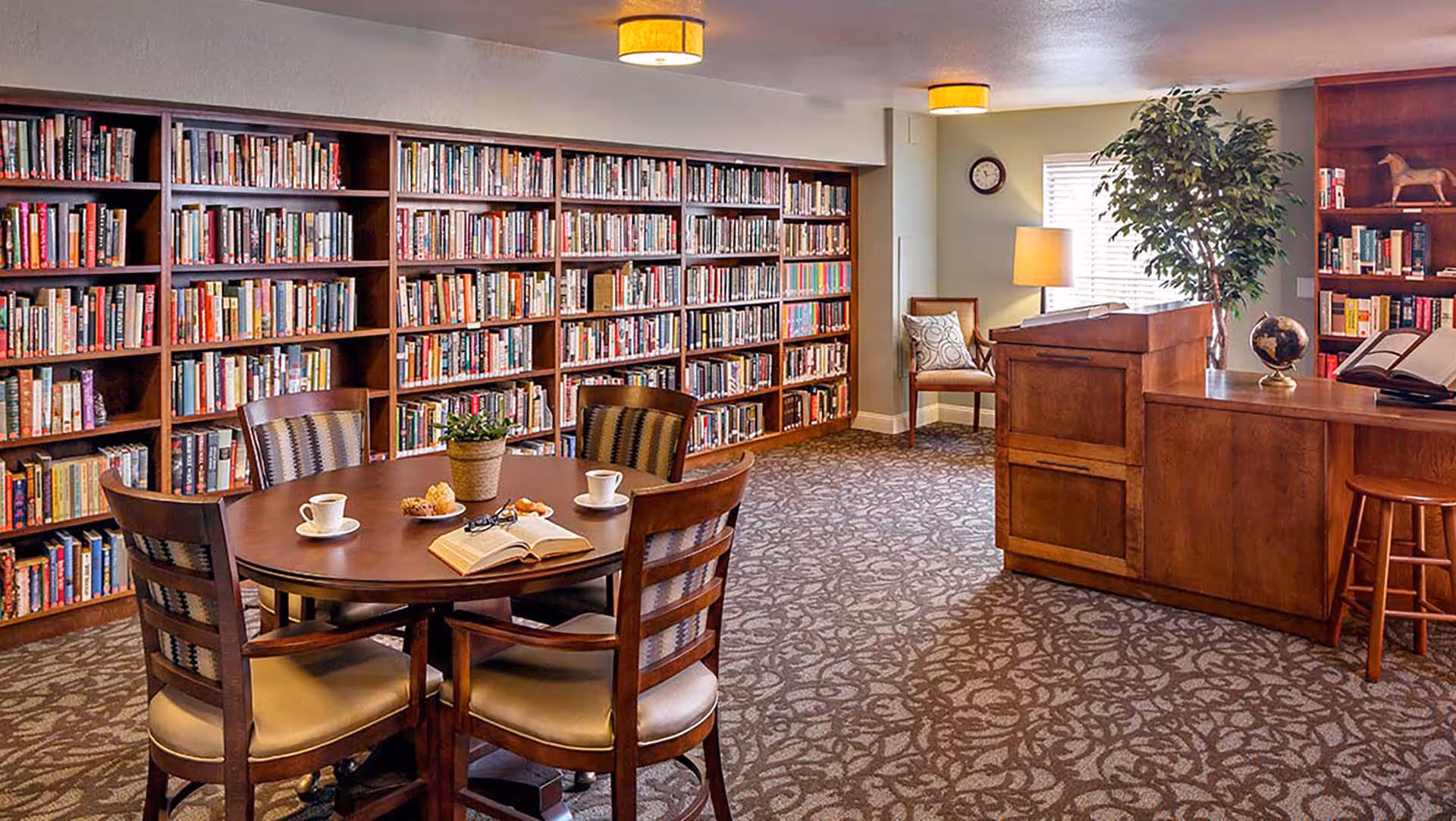 A cozy library room with wooden bookshelves filled with books along the wall, a round wooden table with four chairs, a potted plant, cups and pastries on the table, a wooden desk with a globe and an open book, a chair with a cushion near a window, and warm ceiling lights.