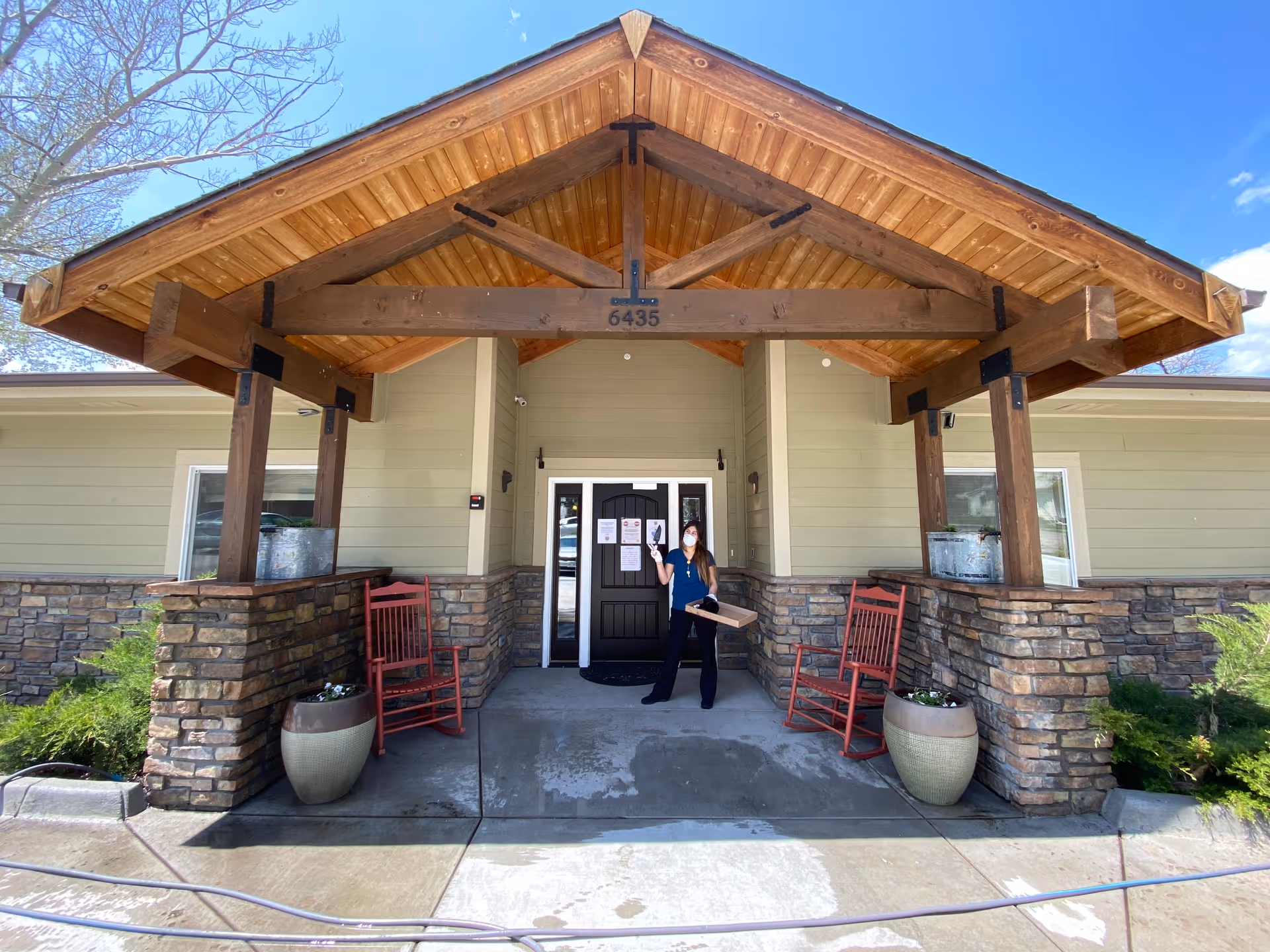 Front entrance of a single-story building with a large wooden porte-cochere, stone columns, red rocking chairs, planters, and a masked person holding a box at the door.