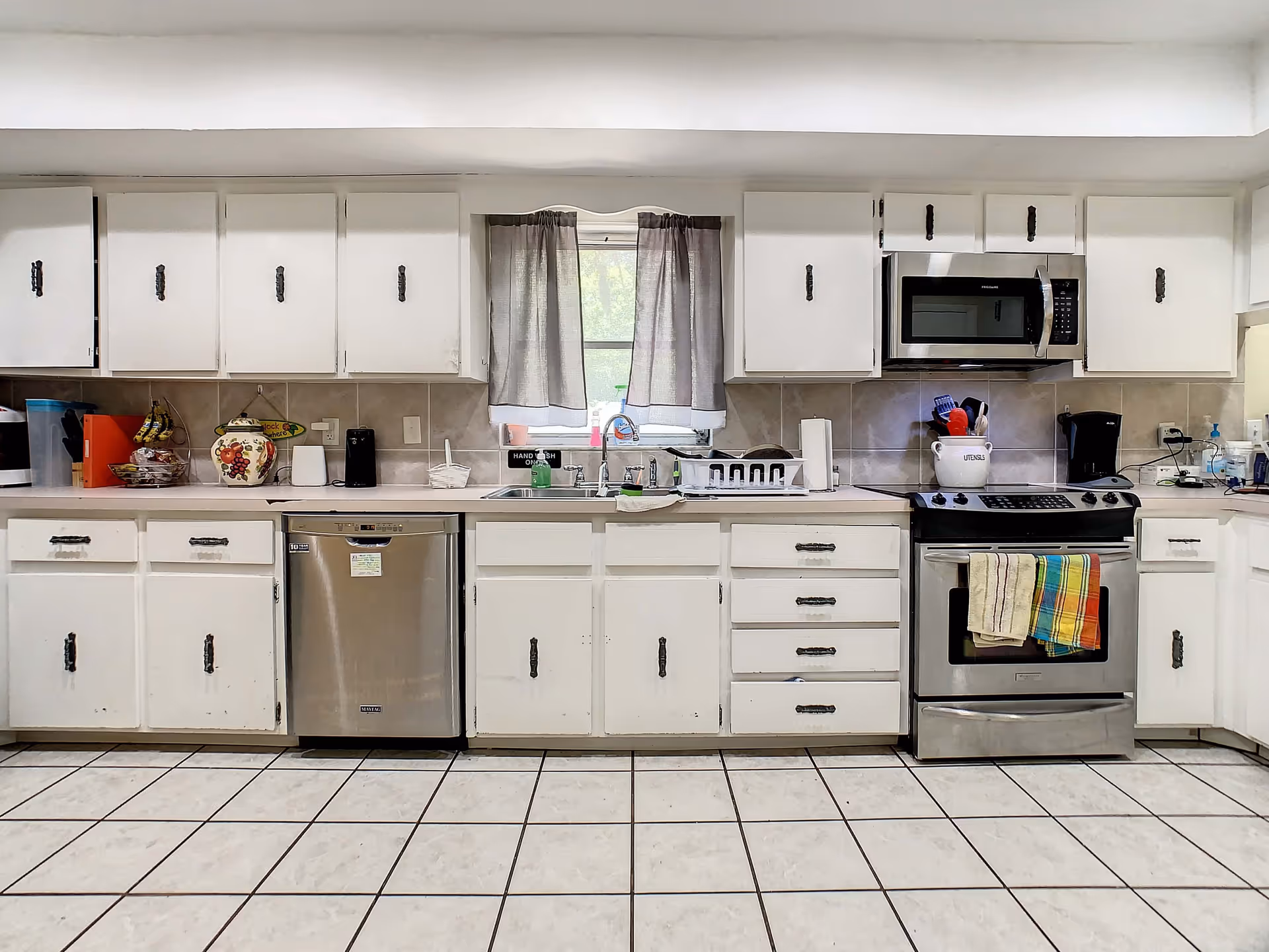 A clean kitchen with white cabinets and drawers, a stainless steel dishwasher, stove, and microwave. There is a window with gray curtains above the sink, and various kitchen items such as a utensil holder, coffee maker, and dish rack on the countertops. The floor is tiled with white tiles and dark grout lines.