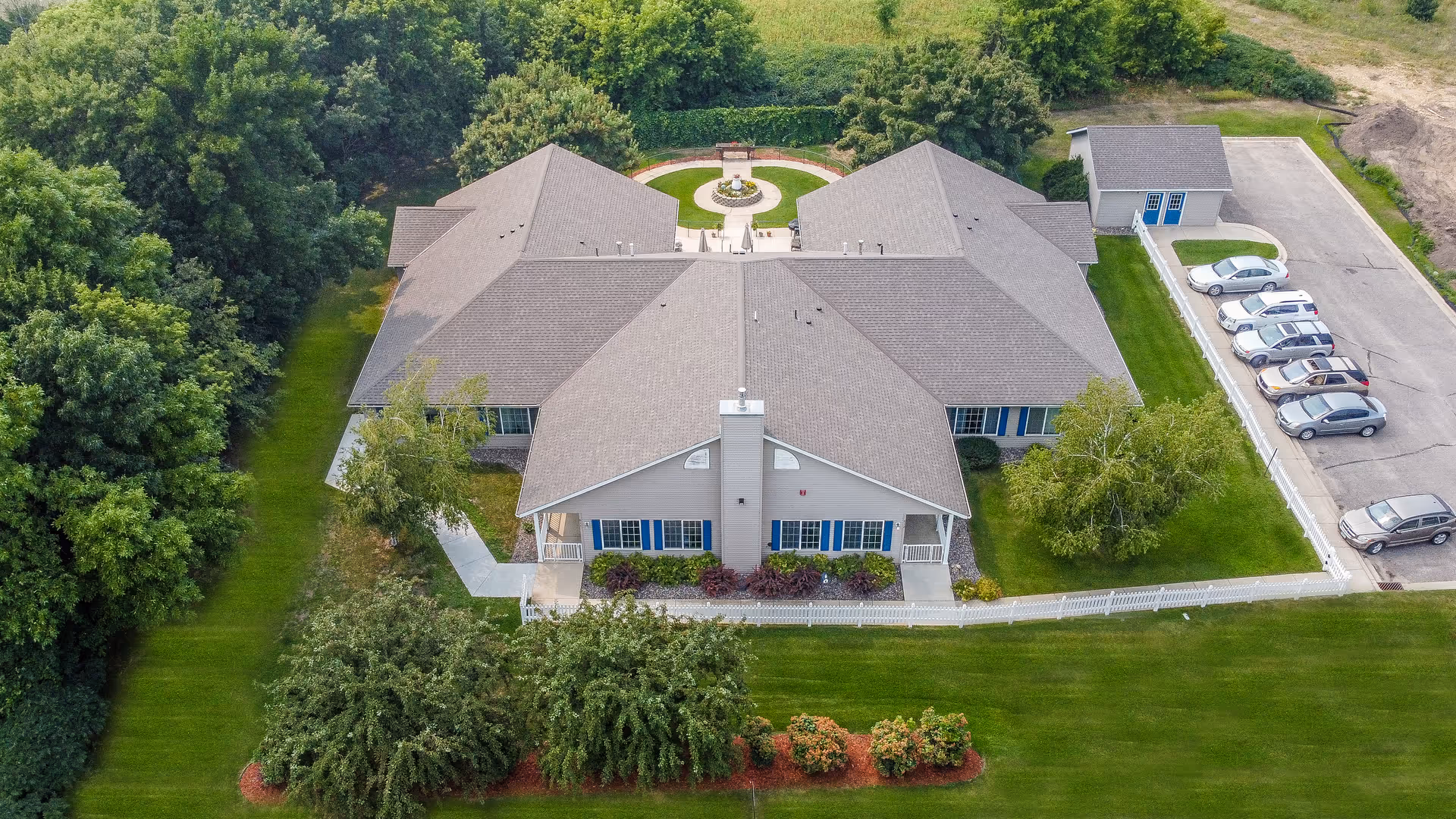 Aerial view of Autumn Cottages by Vivie, showing a large single-story building with a gray roof surrounded by green lawns, trees, and a white fence. There is a circular garden feature behind the building and a parking area with several cars to the right.