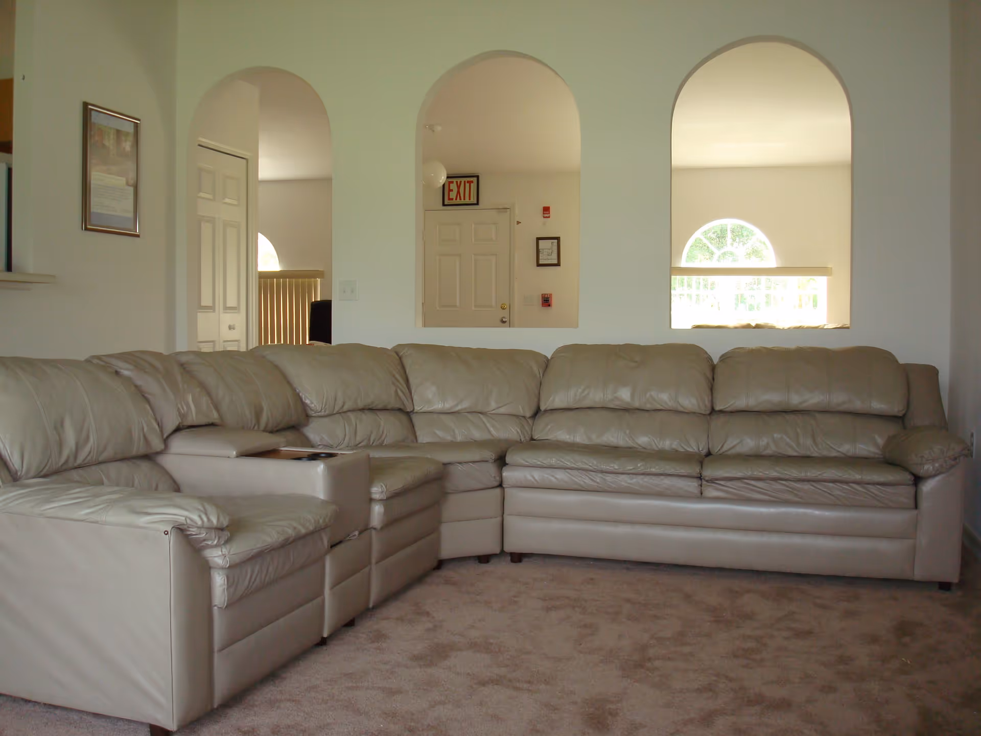 A beige leather sectional sofa in a living room with carpeted floor. The wall behind the sofa has three arched openings showing a door with an exit sign and windows letting in natural light.