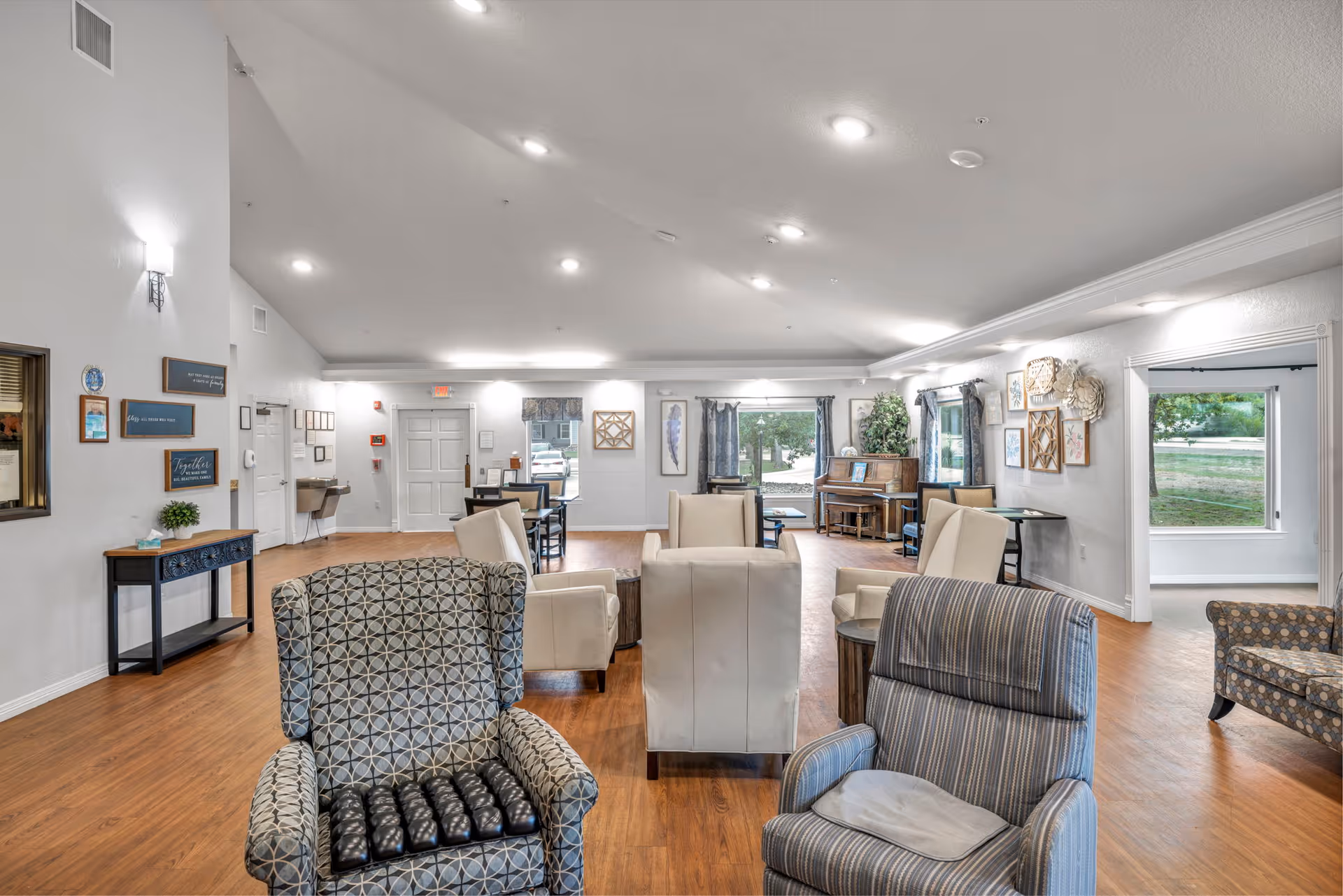 Spacious senior living common room with multiple armchairs and seating areas, a piano, and large windows over wood floors.
