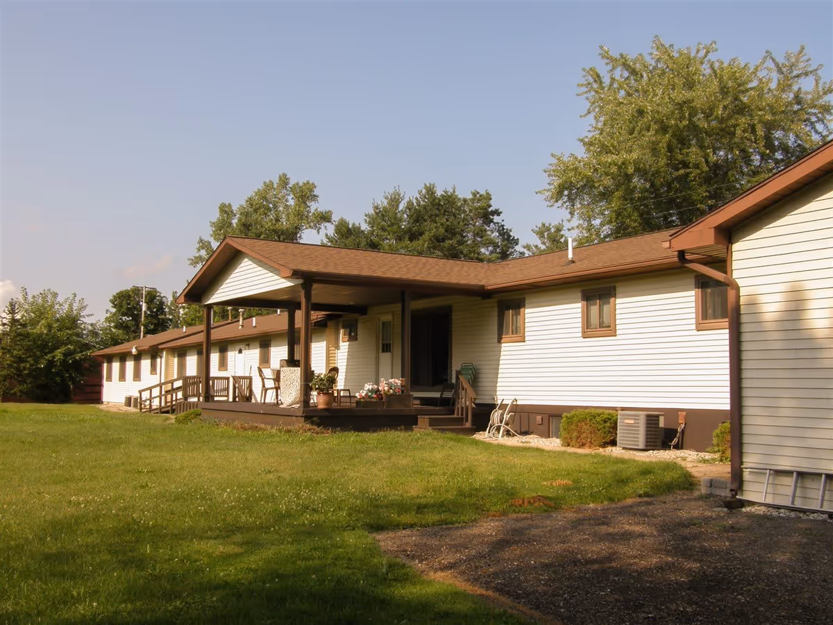 Exterior view of a single-story building with white siding and brown trim, featuring a covered porch with chairs and potted plants. The building is surrounded by green grass and trees under a clear sky.
