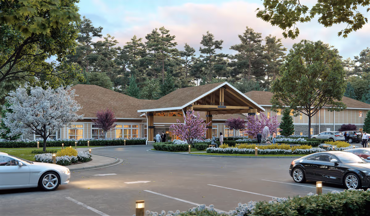 Front entrance of a senior living building with a covered porte-cochère, landscaped roundabout, trees and parked cars.