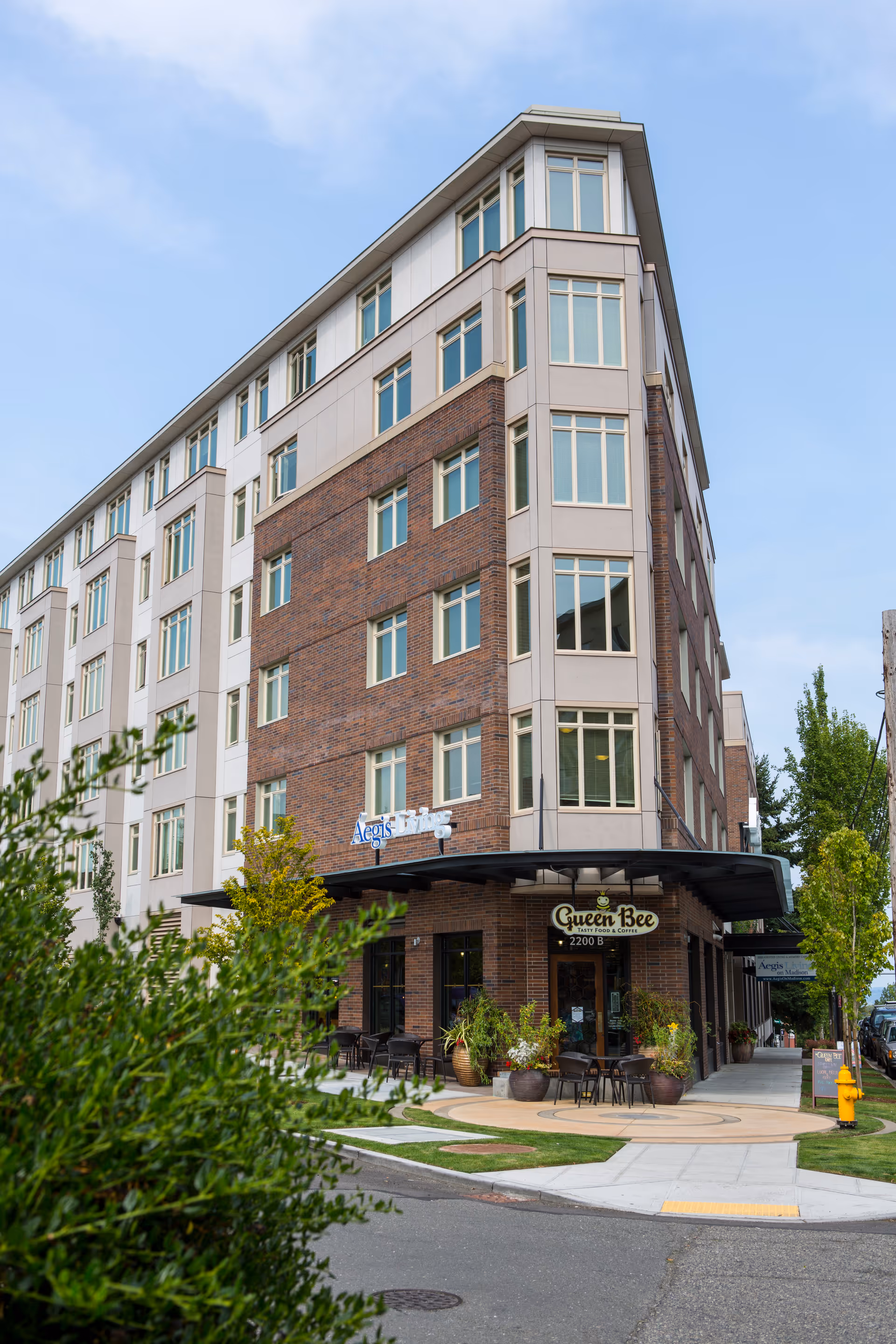Exterior view of a multi-story building with a brick and beige facade. The building has many windows and a sign that reads 'Aegis Living'. There is a cafe named 'Queen Bee Tasty Food & Coffee' on the ground floor with outdoor seating and potted plants. The sky is clear and blue.