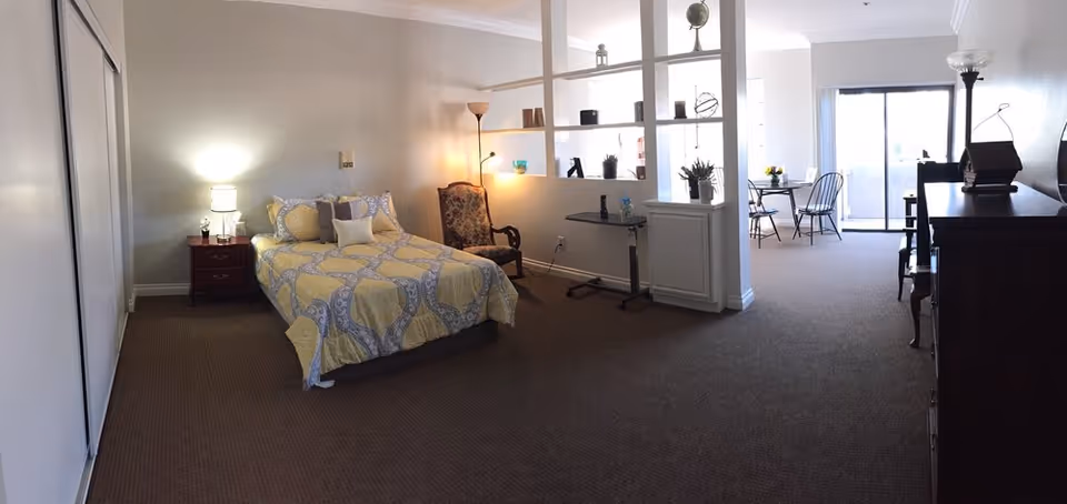 A spacious room in a senior living facility with a bed covered in a yellow and gray patterned comforter, a wooden nightstand with a lamp, an upholstered armchair, a small table, and a shelving divider separating the sleeping area from a dining area with a table and chairs near a sliding glass door leading outside.