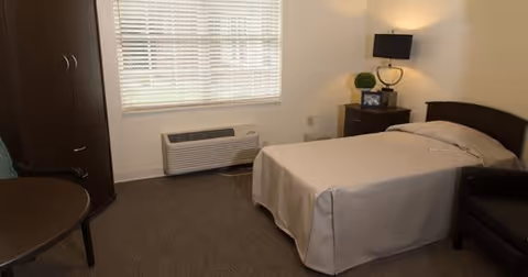 A simple bedroom with a single bed covered in a beige bedspread, a dark wooden nightstand with a lamp and decorative items, a dark wooden wardrobe, a round table with a chair, a window with blinds, and a wall-mounted air conditioning unit below the window.