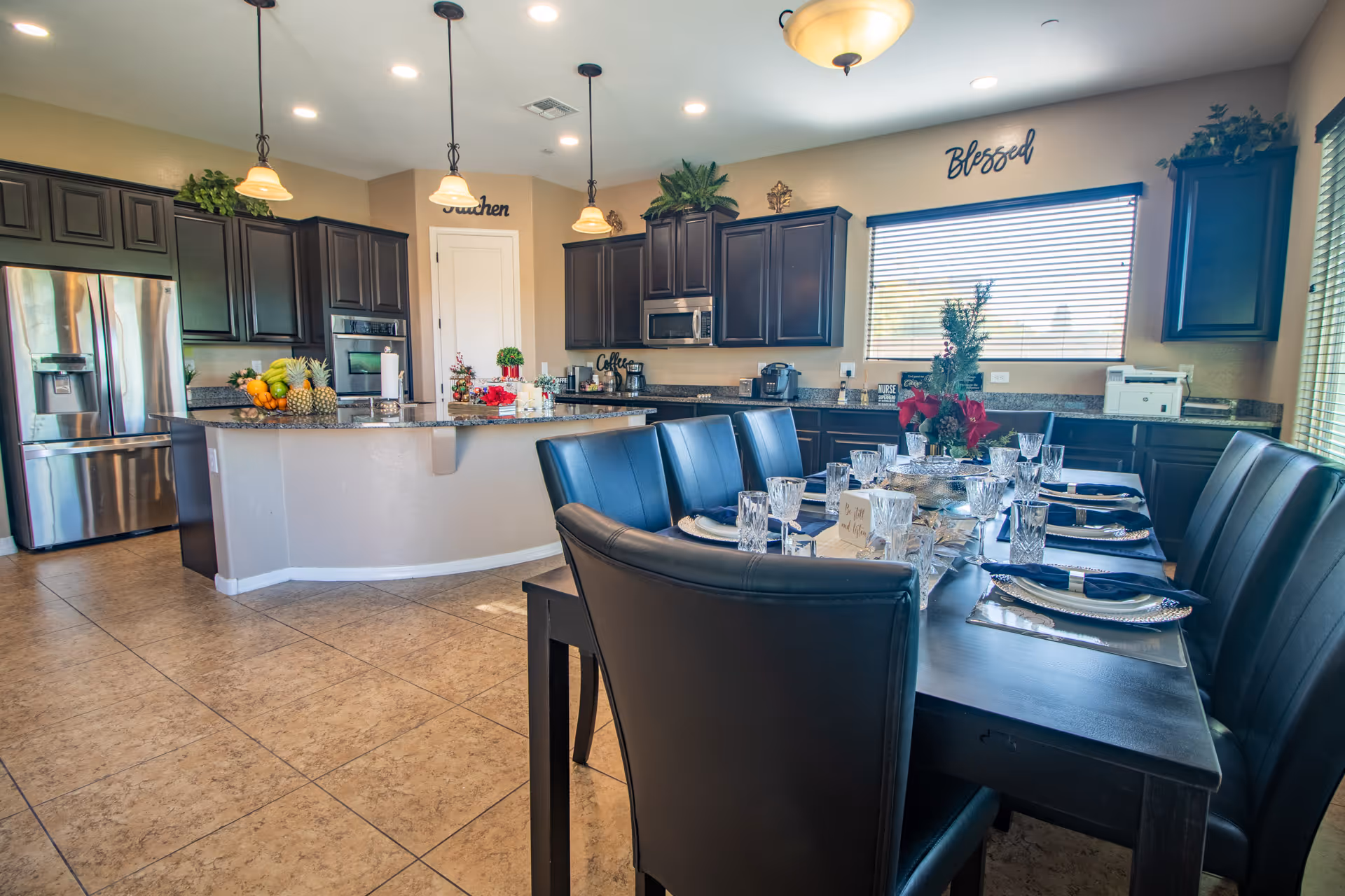 Open-plan dining area and kitchen with a large set dining table, dark cabinets, stainless appliances and a center island under pendant lights.