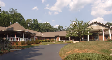Exterior view of a single-story senior living facility building with a covered entrance and a wraparound porch, surrounded by greenery and trees under a partly cloudy sky.