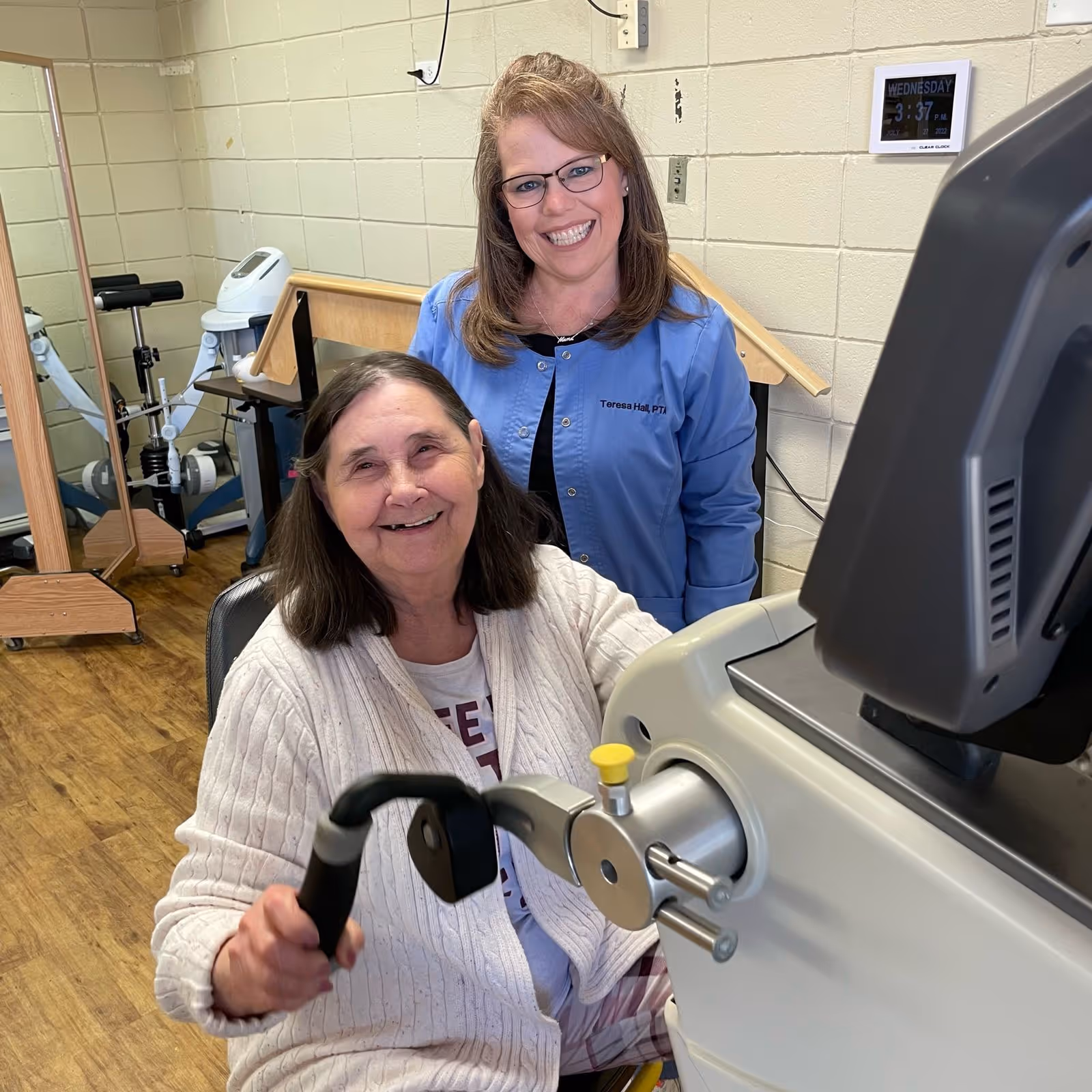 An elderly woman using a hand exercise machine in a therapy room, smiling, with a female physical therapist standing behind her also smiling. The room has wooden flooring, a large mirror, and various therapy equipment.