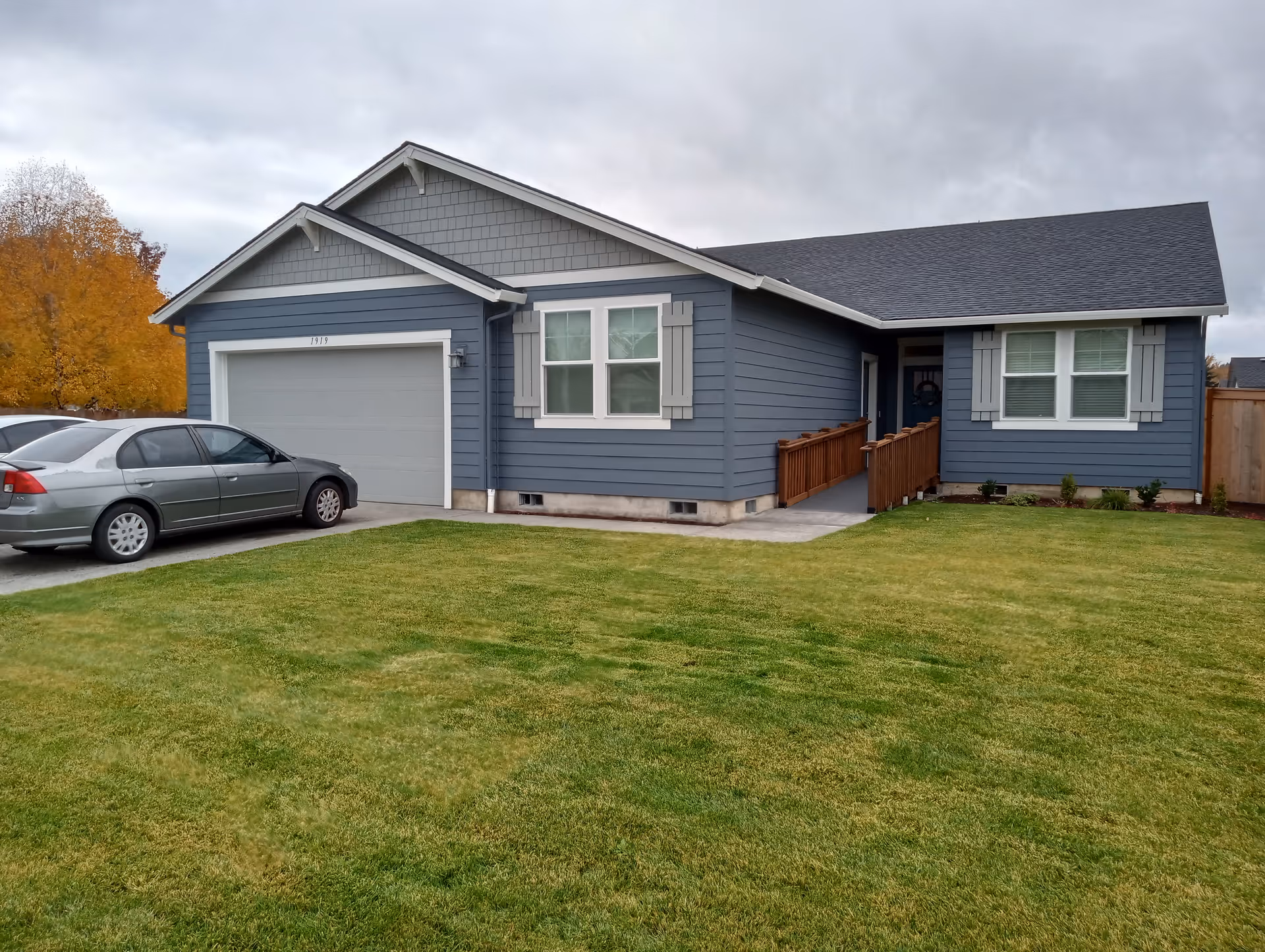 Exterior view of a single-story blue house with white trim, a two-car garage, a wooden ramp leading to the front door, and a well-maintained green lawn. Two cars are parked in the driveway, and there are autumn-colored trees in the background under a cloudy sky.