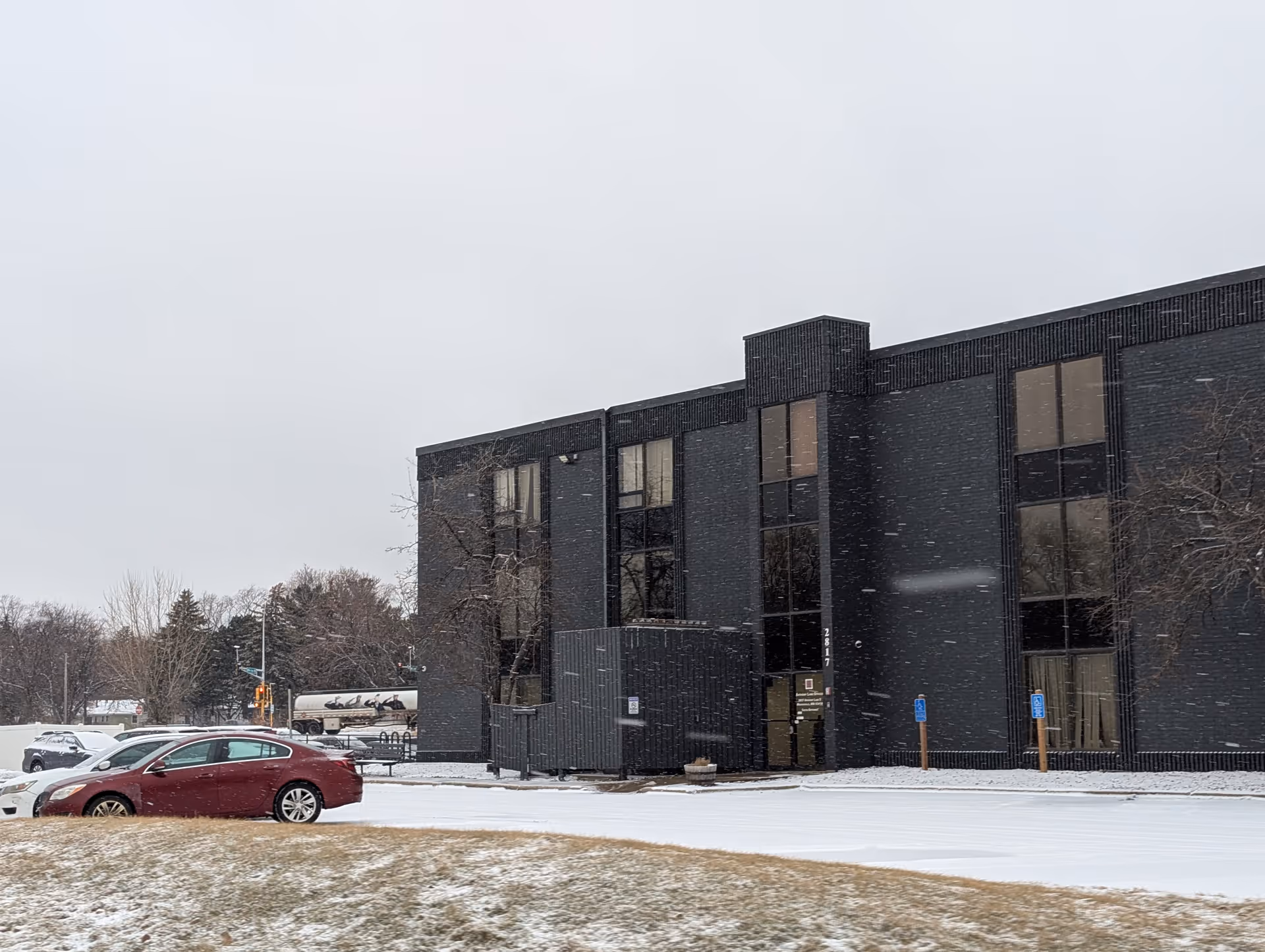 Exterior view of a modern two-story dark brick building with large windows, surrounded by a snowy landscape and a few parked cars in the parking lot.