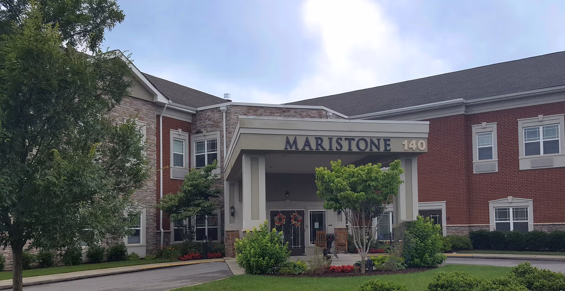 Exterior front entrance of Maristone of Providence, a two-story brick building with a covered porch area, surrounded by trees and landscaping under a cloudy sky.