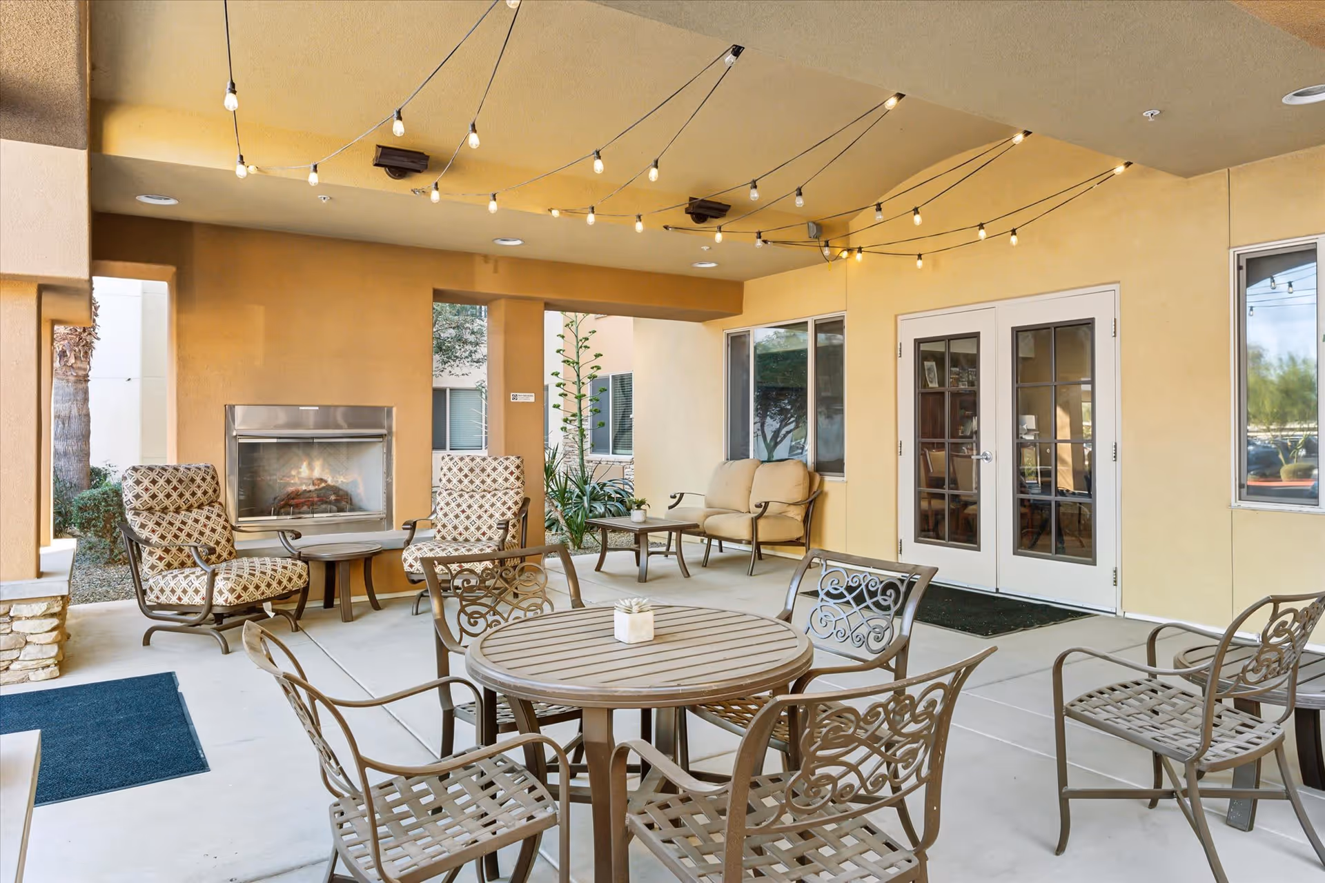 Covered outdoor patio area with string lights hanging from the ceiling, a round metal table with four matching chairs, two cushioned armchairs near a built-in fireplace, and a cushioned loveseat with a small coffee table. The walls are beige, and there are glass double doors and windows leading inside.