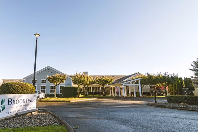 Exterior view of a senior living facility building with a driveway, small trees, and a sign that reads 'Brookdale Ocean Shores Senior Living Solutions' under a clear blue sky.