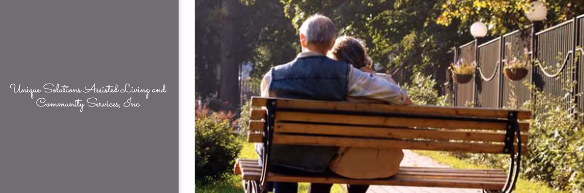 An elderly couple sitting closely together on a wooden bench in a sunlit outdoor garden area, surrounded by greenery and hanging flower pots along a fence.