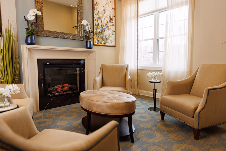 A cozy common room with beige armchairs arranged around a round ottoman in front of a fireplace and a sunlit window.