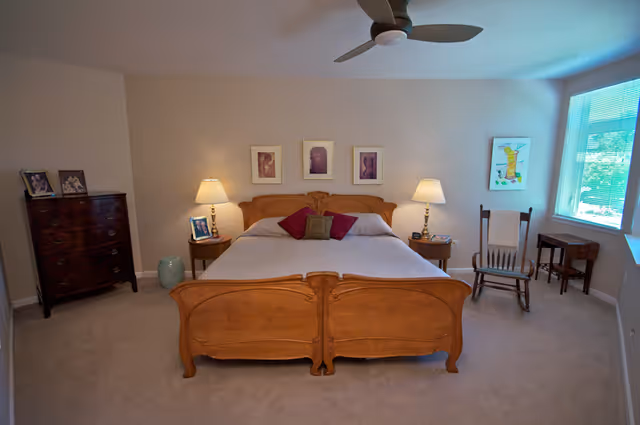 A bedroom with a wooden double bed featuring a carved headboard and footboard, two bedside tables each with a lamp, a wooden rocking chair near a window with blinds, a small table, and a dark wooden dresser with framed photos on top. The walls are light-colored and there are three framed pictures above the bed.