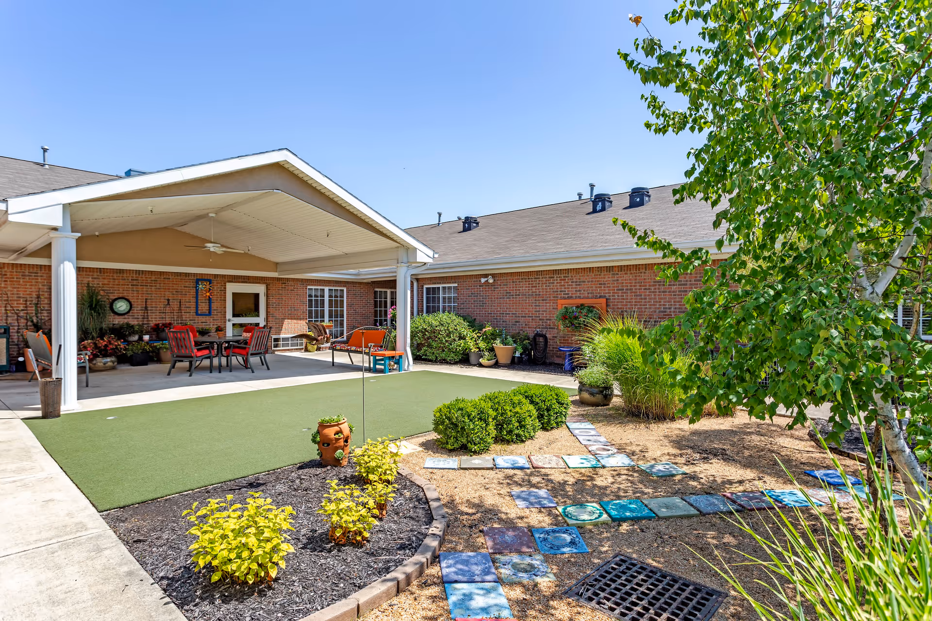 Outdoor courtyard area at Morrison Woods Health Campus featuring a covered patio with seating including tables and chairs, surrounded by brick buildings. The courtyard has a green artificial turf area, landscaped garden beds with plants and bushes, and a decorative tiled pathway. The sky is clear and blue.