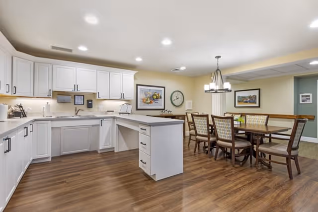 A bright and clean kitchen and dining area with white cabinets, a countertop island, wooden flooring, and a dining table with six chairs. The walls are decorated with framed artwork and a clock, and there is a modern light fixture hanging above the dining table.