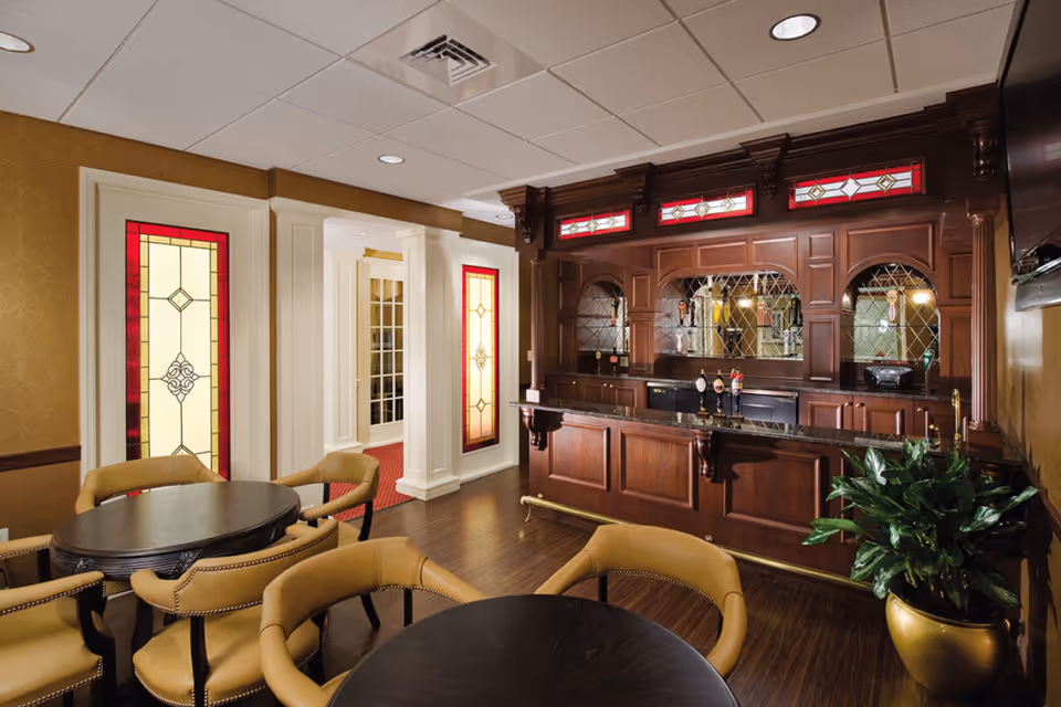 Interior view of a lounge area with a dark wooden bar featuring stained glass windows above it. There are round tables with tan upholstered chairs arranged around the room. The walls have decorative stained glass panels and the floor is dark wood. A potted plant is placed near the bar.