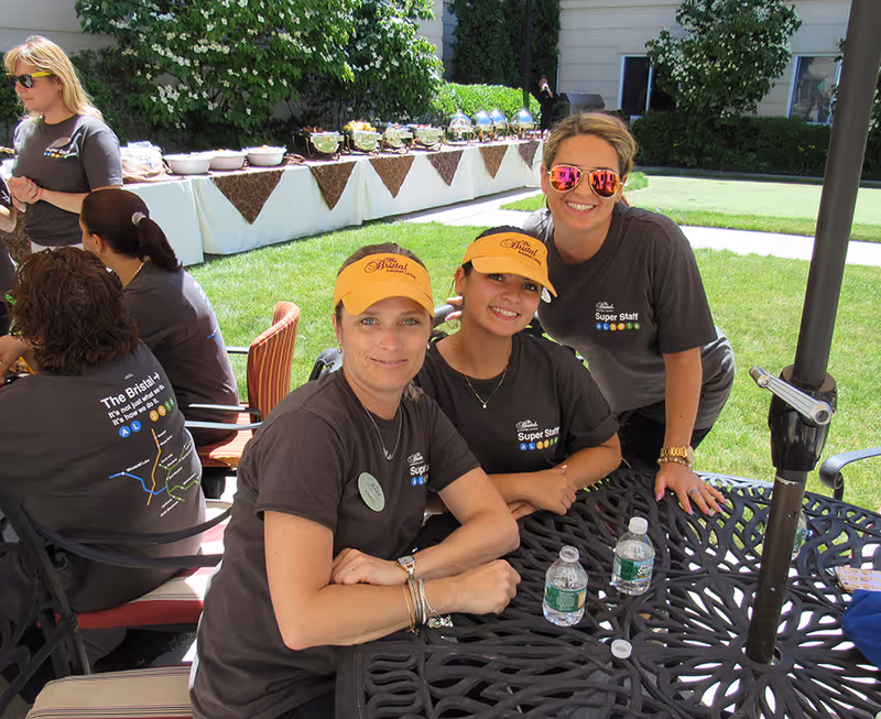 Three women wearing The Bristal Assisted Living t-shirts and yellow visors sitting at a black metal outdoor table with water bottles, smiling at the camera. In the background, there is a long buffet table with food covered by chafing dishes and bowls, set up on a lawn outside a building.