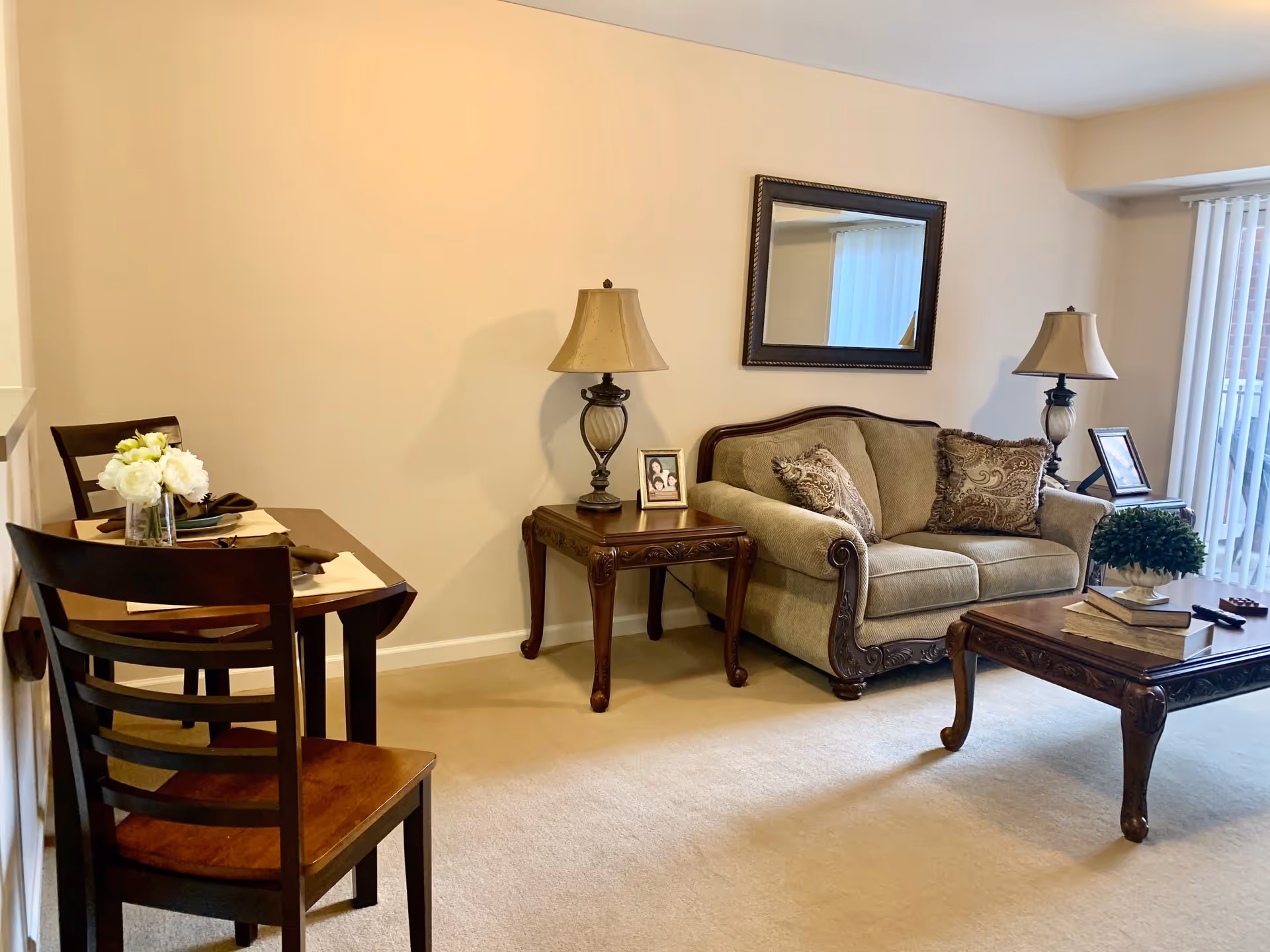 A cozy living room with a beige couch adorned with patterned pillows, two wooden side tables each with a lamp, a wooden coffee table with books and a small plant, a wooden dining table with two chairs and a vase of white flowers, and a large mirror on the wall reflecting a window with vertical blinds.