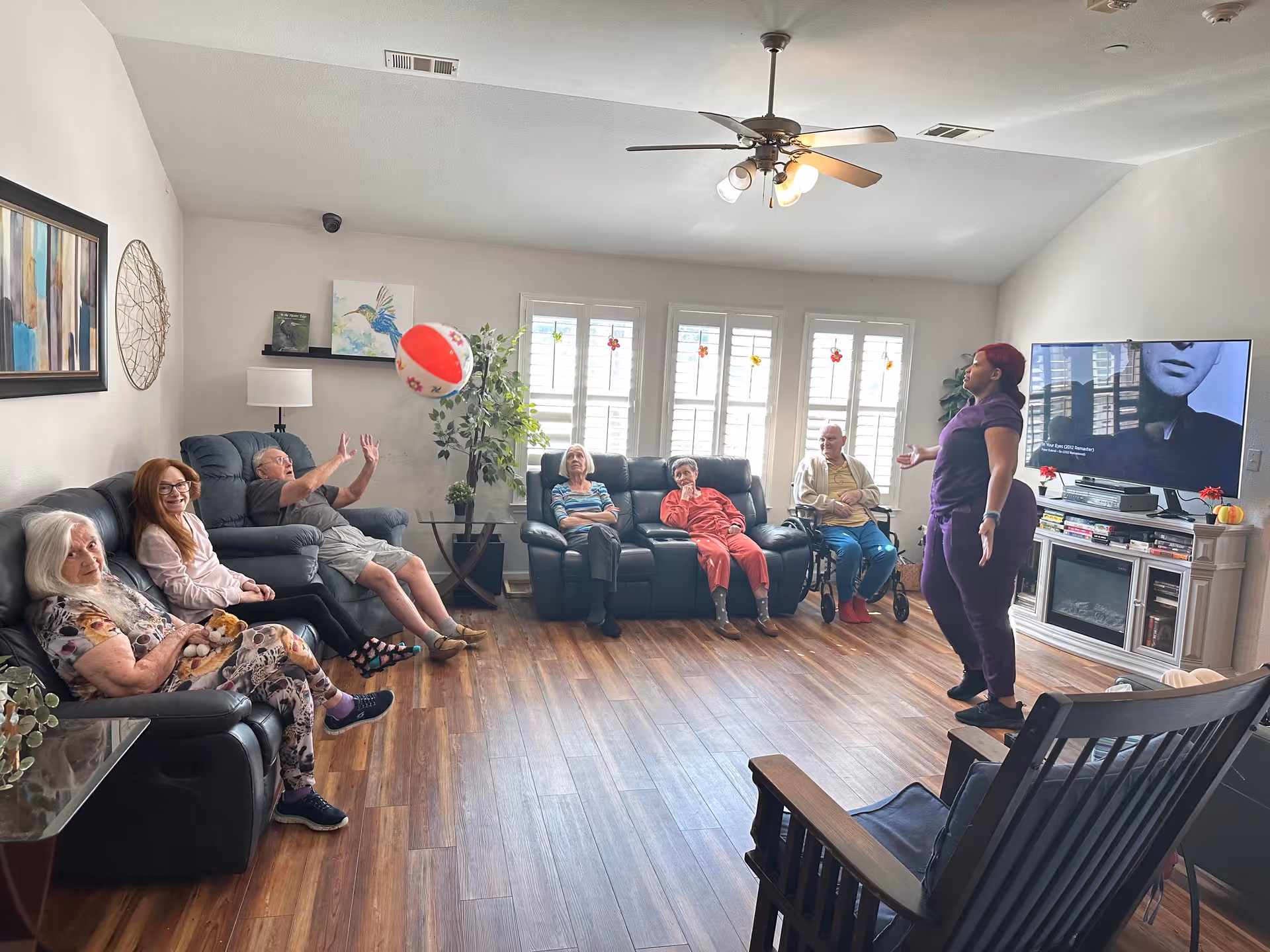 A group of elderly people sitting on sofas in a living room playing with a beach ball, while a caregiver stands nearby engaging with them. The room has wooden flooring, a ceiling fan, a TV on a stand, and large windows with white shutters letting in natural light.