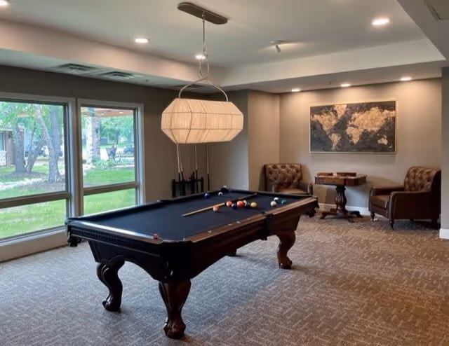 A well-lit recreational room with a black felt pool table in the center, a modern hanging light fixture above it, two brown leather armchairs, a small wooden table, and a world map artwork on the wall. Large windows show a green outdoor area with trees.
